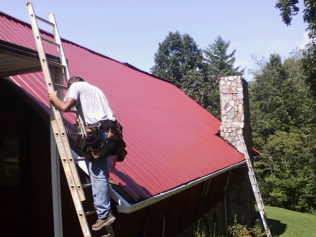 Roofer on a ladder beside a red metal roof and stone chimney — Soffit & Fascia Repair by Roofing Brothers in Pomona